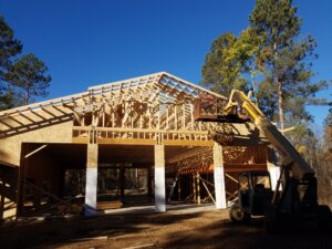 A partially constructed wooden building with visible framing, surrounded by tall trees. A yellow forklift raises a worker toward the roof trusses under a clear blue sky.