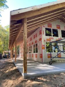 A house under construction with exposed wooden beams, partially installed windows, and insulation wrap on the exterior walls. Construction equipment and scaffolding are visible near the building.