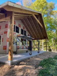 A house under construction with exposed wooden beams and scaffolding on a concrete porch, surrounded by trees and dirt, under a clear blue sky.