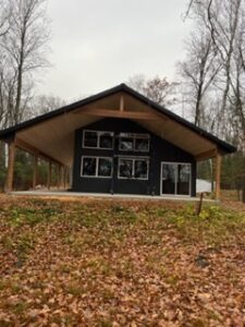 A modern cabin with large windows and a wide overhanging roof stands in a wooded area. Fallen autumn leaves cover the ground, and bare trees surround the house under an overcast sky.
