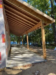 A partially constructed wooden porch with a slanted roof extends alongside a building near a wooded area, with a lake visible in the background on a sunny day.