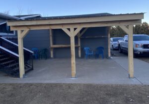 A wooden patio cover with a sloped roof stands on a concrete slab next to a gray house. Three blue plastic chairs are arranged underneath, and two pickup trucks are parked nearby. Stairs are visible on the left.