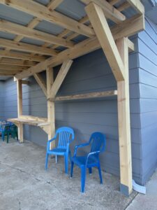 Two blue plastic chairs sit under a wooden pergola attached to a gray-sided building, with a concrete floor and additional green and blue chairs visible in the background.