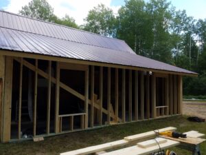 A partially constructed wooden building with a metal roof, showing exposed wall framing and beams. Tools and lumber are on the grass nearby, with trees and a cloudy sky in the background.