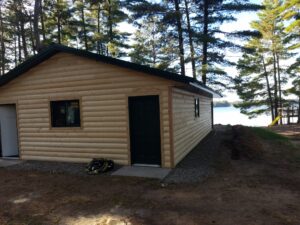 A log cabin with a dark green door and window sits among tall pine trees near a lake, with sunlight shining through the trees and a coiled hose on the ground beside the cabin.