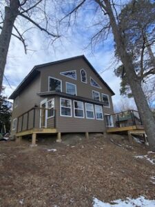 A two-story house with gray siding and multiple large windows sits on a sloped, leaf-covered yard with a few patches of snow and tall trees, under a partly cloudy sky.