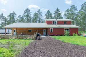 A large two-story house with wood and red siding, a metal roof, fenced garden beds, a grill on a mulched path, and tall pine trees in the background under a blue sky.