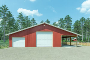 A large red metal building with three white garage doors, one of which is open, stands on a dirt lot surrounded by tall pine trees under a bright blue sky.