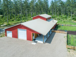 Aerial view of a large red barn-style building with a light-colored metal roof, set in a fenced rural area surrounded by tall pine trees and a small garden plot.