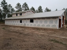 A large, partially constructed building with a white metal roof and walls covered in Tyvek house wrap sits on bare dirt, surrounded by pine trees under an overcast sky. Some windows and door frames are visible.