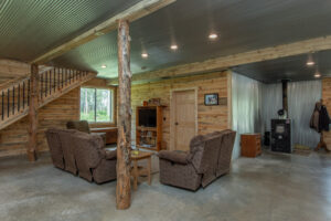 A cozy living room with rustic wood walls, exposed beams, brown upholstered furniture, a wood stove in the corner, and a staircase leading up, all set on a concrete floor with natural light coming through the window.
