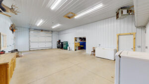 A spacious, well-lit garage with white paneled walls and ceiling, concrete floor, workbenches on the left, toolboxes and shelves at the back, a freezer on the right, and a closed double garage door.