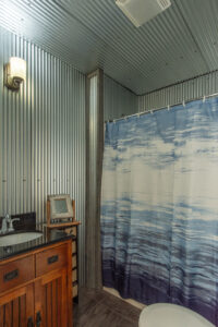 A rustic bathroom with corrugated metal walls and ceiling, a wooden vanity with a sink, a small shelf, and a blue and white shower curtain with an abstract pattern.