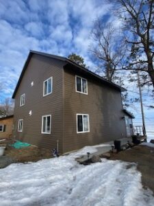 A two-story brown house with white-trimmed windows stands on a snowy yard under a partly cloudy blue sky. Leafless trees are nearby, and another building is partially visible on the left.