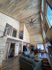A cozy cabin interior with high vaulted wooden ceilings, large windows, a loft area, kitchen, dining table, and a living space furnished with a sofa and chairs on a colorful rug.