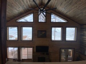 A large, wooden cabin interior with tall windows offering views of a snowy, lakeside landscape. The high ceiling has exposed wood and a modern black fan. A TV sits above a stone fireplace.