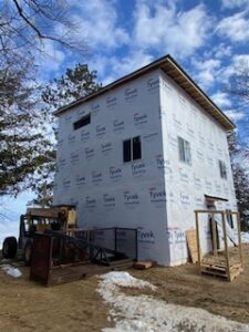 A two-story building under construction, wrapped in Tyvek house wrap, stands on a dirt lot with patches of snow. A forklift and building materials are nearby under a partly cloudy sky.