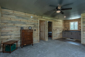 A rustic room with light wood log walls, carpeted floor, a wooden dresser, small table with crates, ceiling fan, and a separate area with a sink and cabinets. A doorway leads to a bedroom. Natural light from windows.