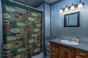 A rustic bathroom with corrugated metal walls, a wooden vanity, a framed mirror, and a green shower curtain decorated with cabin and nature-themed signs. Four bulbs light up the mirror above the sink.