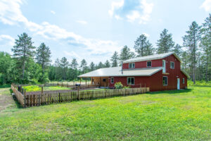 A large red barn-style house with a metal roof sits on a grassy lawn, next to a fenced garden area, surrounded by tall pine trees under a partly cloudy sky.
