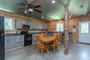 A rustic kitchen with light wood walls, concrete floors, gray cabinets, black appliances, a round wooden dining table with four chairs, a ceiling fan, and a support post made from a natural tree trunk.