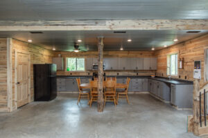Spacious rustic kitchen with wood-paneled walls, gray cabinets, a black refrigerator, a central wooden dining table with six chairs, concrete floors, and exposed wooden beams. Natural light enters through two windows.