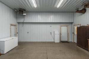 A clean, spacious garage with metal walls and ceiling, concrete floor, a chest freezer, refrigerator, two white doors, wood trim, mounted deer heads, and ceiling lights.