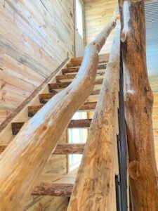 A close-up view of a wooden staircase with rustic, natural log railings inside a building with wooden plank walls and abundant natural light.
