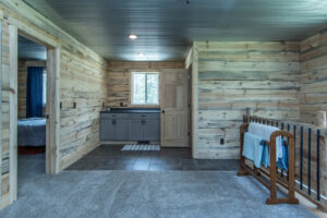 A rustic room with wood-paneled walls and a gray carpet. Theres a wooden crib with towels, a small vanity with cabinets and a window, and an open doorway leading to a bedroom with a bed.