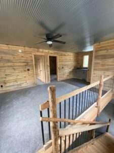 Interior view of a rustic cabin with wood-paneled walls, carpeted floor, a ceiling fan, small kitchenette, and wooden staircase with log railing leading downstairs. Two doorways lead to other rooms.